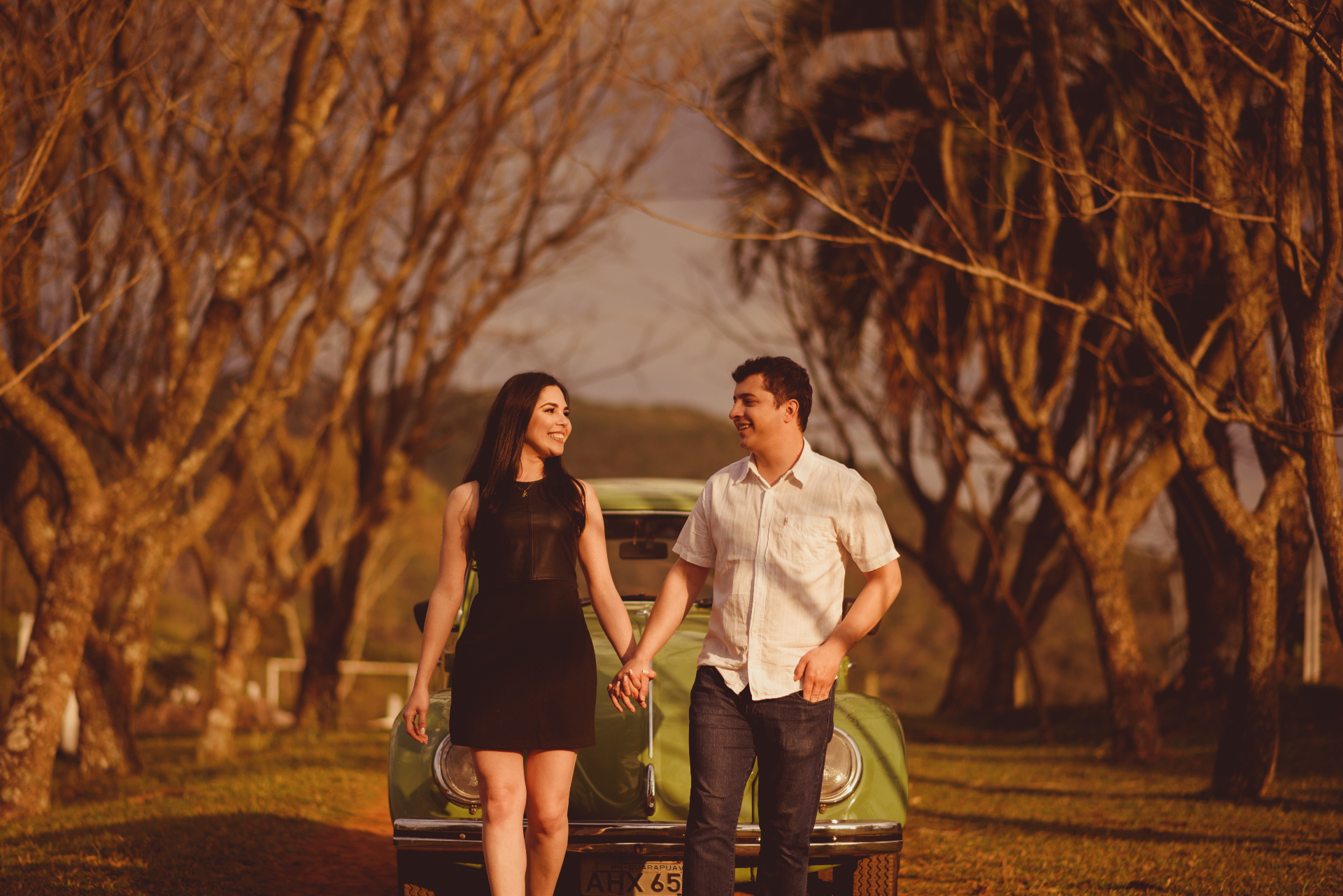 Casal de mãos dadas em frente a Fusca verde sob árvores ao entardecer